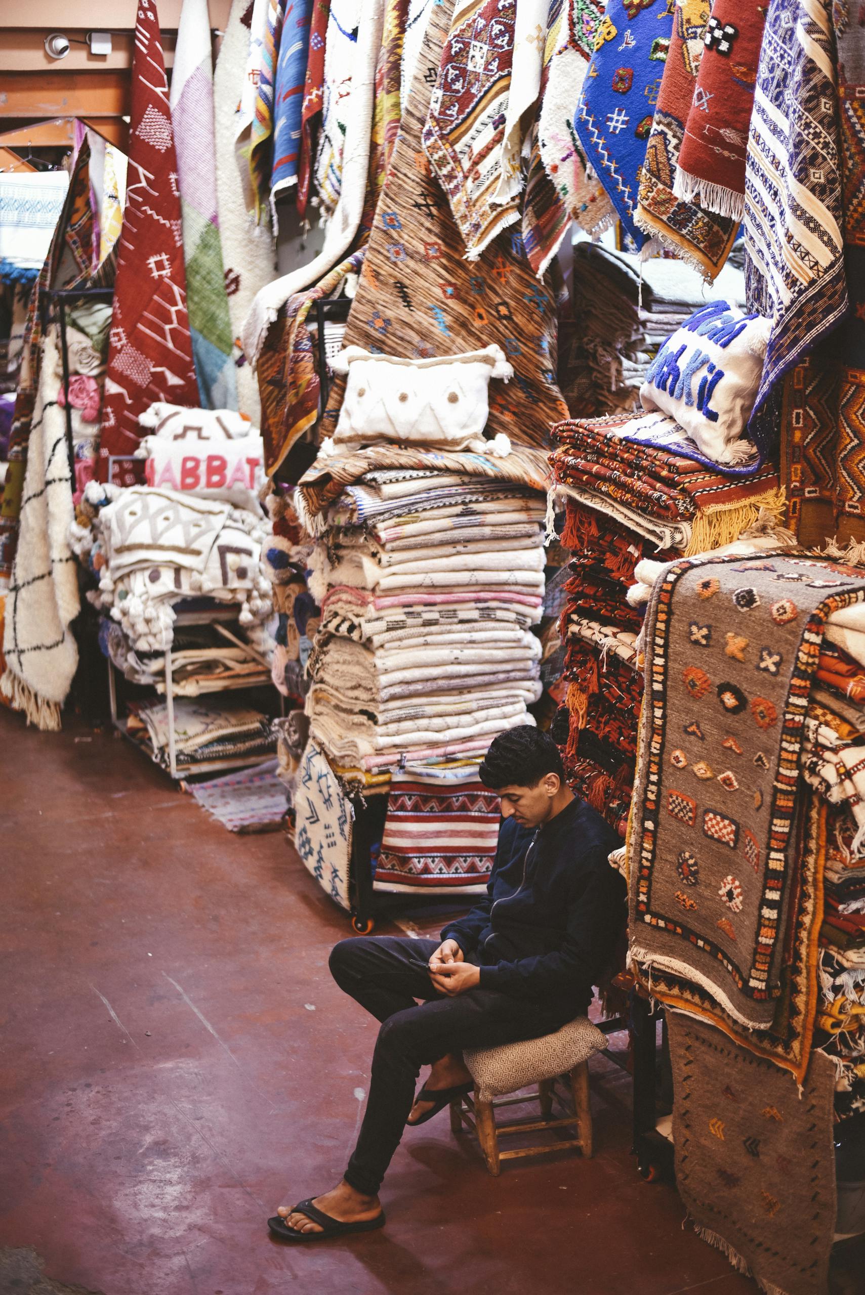 A vibrant display of stacked rugs in a Moroccan market, with a man sitting nearby.